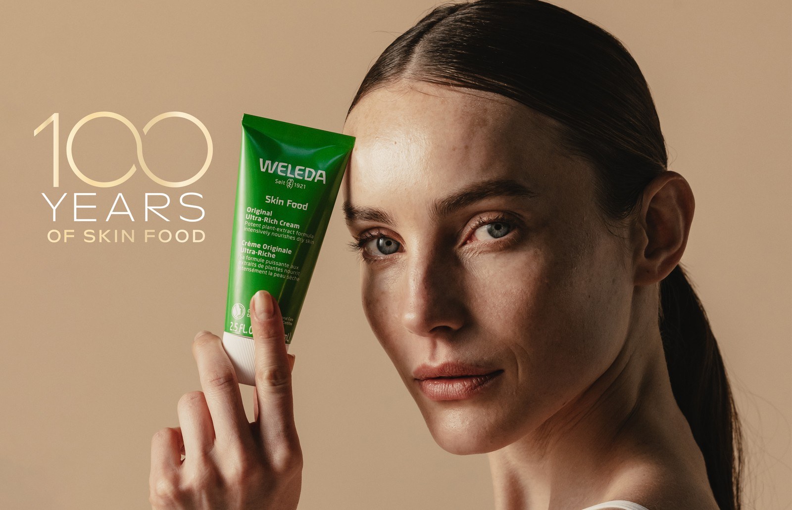 Weleda Skin Food campaign portrait: a model with glowing natural skin holding the green Skin Food tube beside the 100 Years of Skin Food lockup