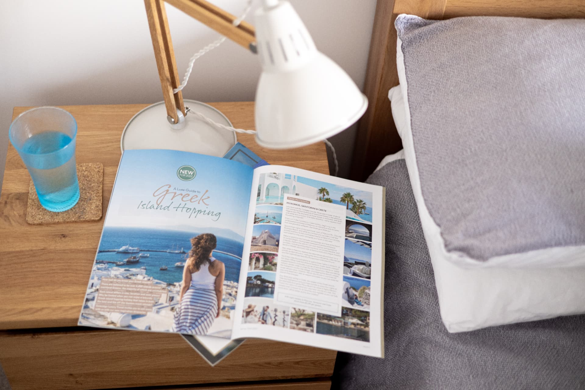 A printed Madhatter travel magazine open on a bedside table beside a lamp and glass of water