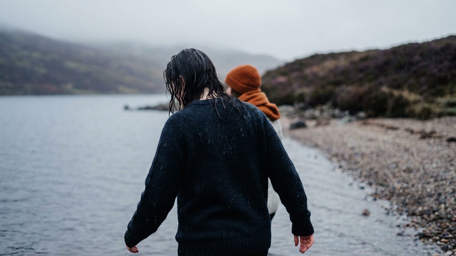 A person exits a cold loch, water dripping, with a misty landscape behind them.